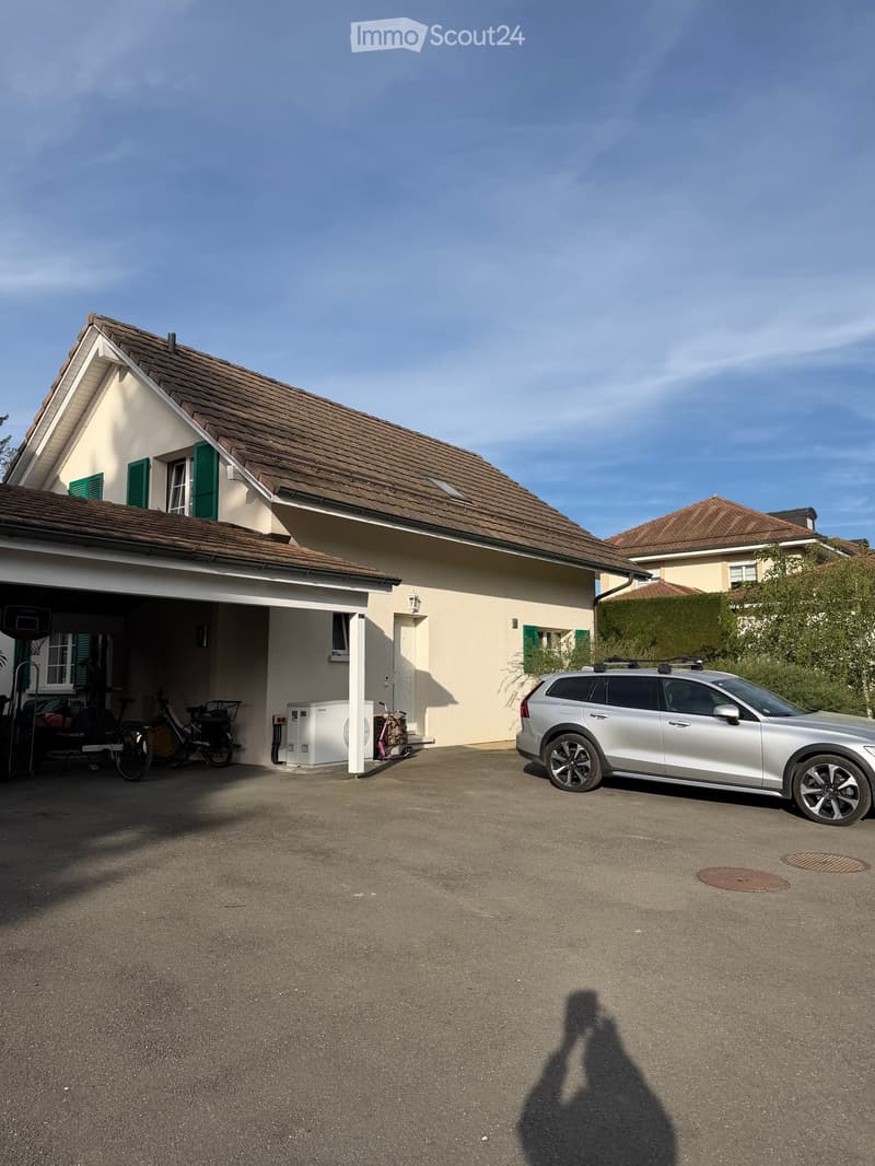 Two-story house with beige walls, green shutters, and a brown tiled roof. A silver SUV is parked in the driveway, and bicycles are stored under the carport.