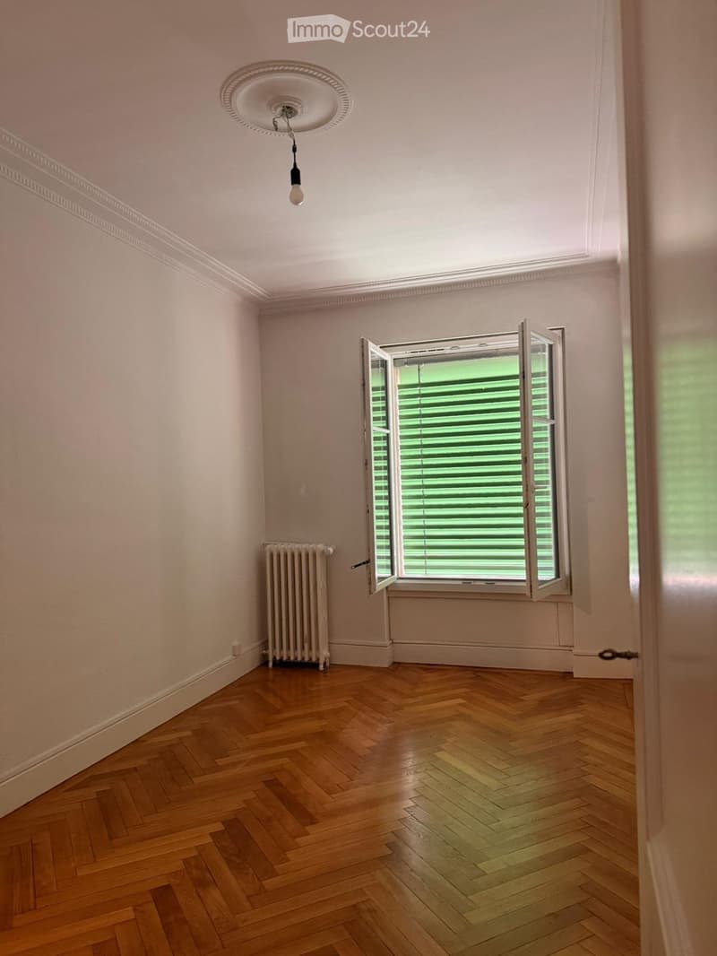 Empty room with wooden herringbone flooring, white walls, a radiator, and a large window with blinds. The ceiling features a decorative light fixture.