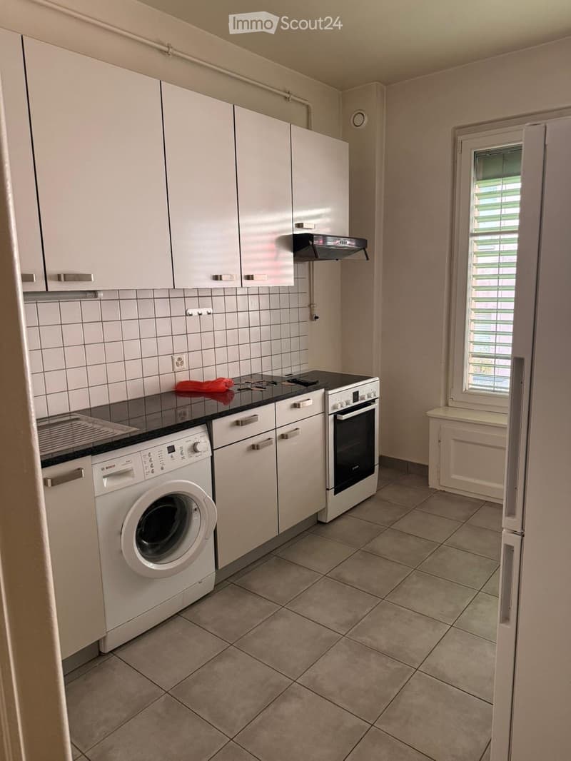 Modern kitchen with white cabinets, a washing machine, an oven, and a stove. The countertops are black, and the floor is tiled.