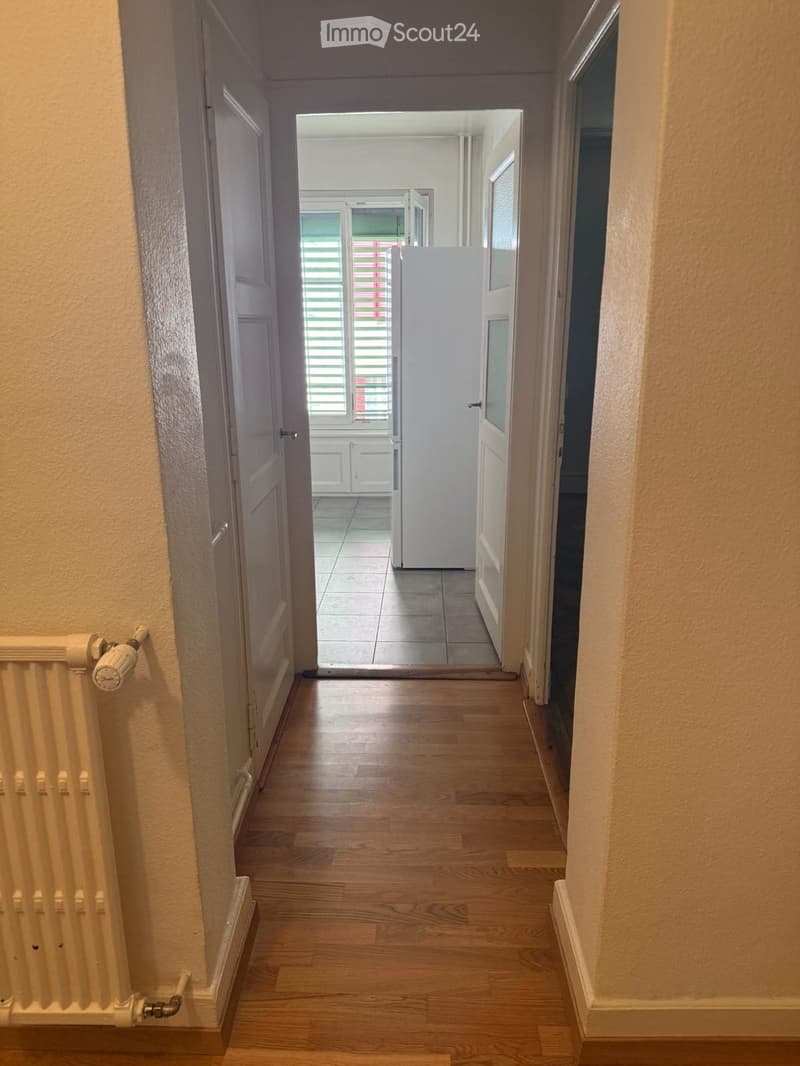 A narrow hallway with wooden flooring, light-colored walls, and a radiator on the left side. The hallway leads to an open doorway revealing a tiled room beyond.