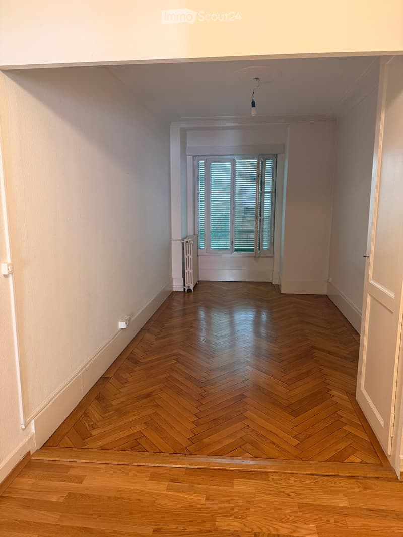 Empty hallway with herringbone wooden flooring, white walls, and a radiator on the left side. A window with shutters is visible at the end of the hallway.