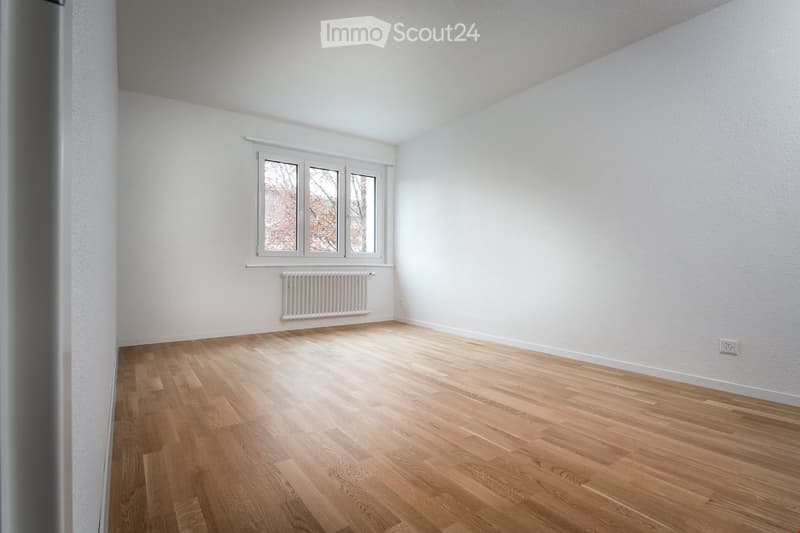 Empty room with wooden floor, white walls, window with radiator, power outlet