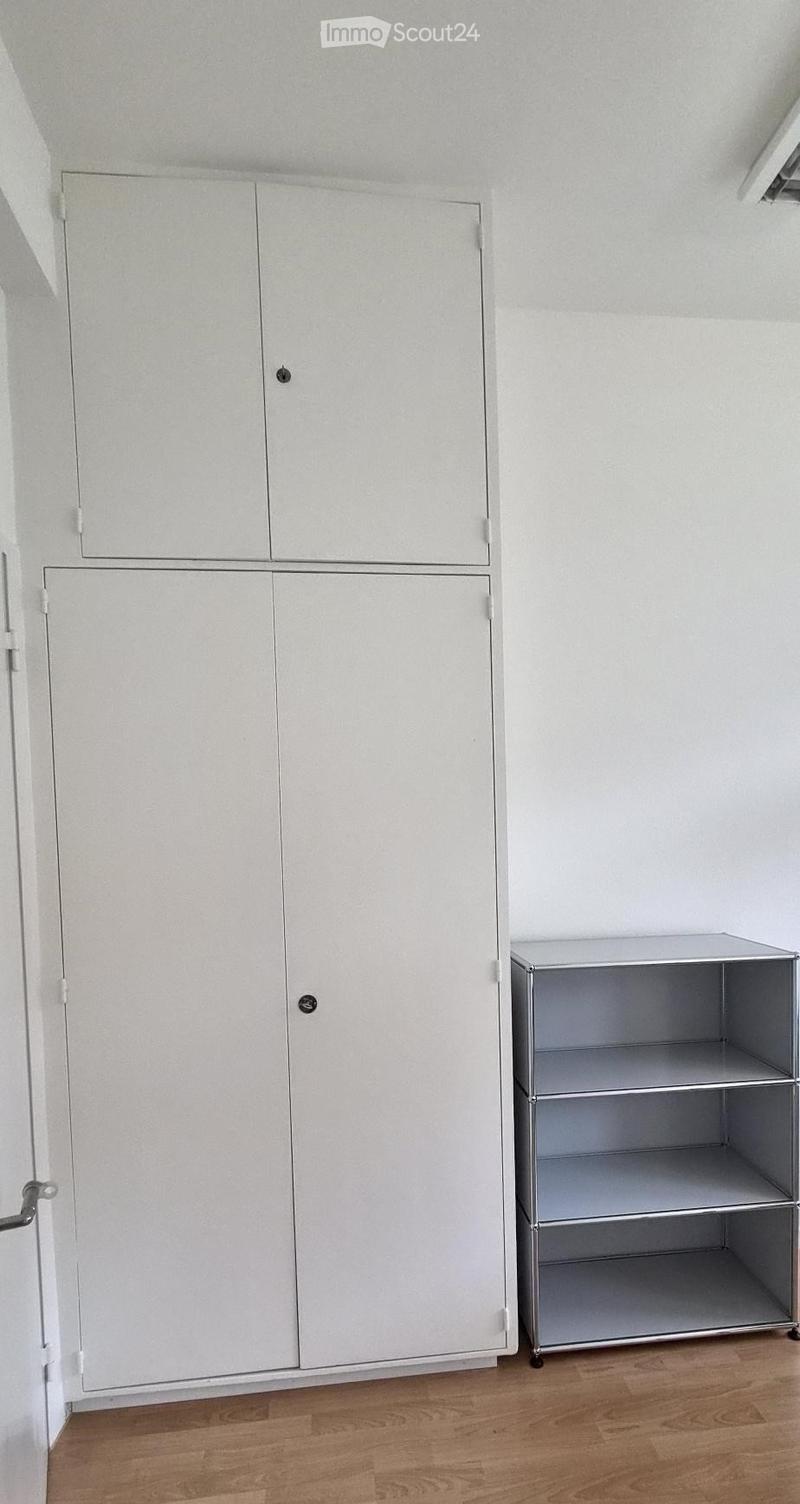 Empty room with light-colored wooden flooring, white walls, and built-in white cabinets with silver handles. A small metal shelving unit is positioned against the wall.