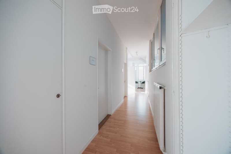 A clean, white-walled hallway with wooden flooring, featuring several doors and a window on the right side.