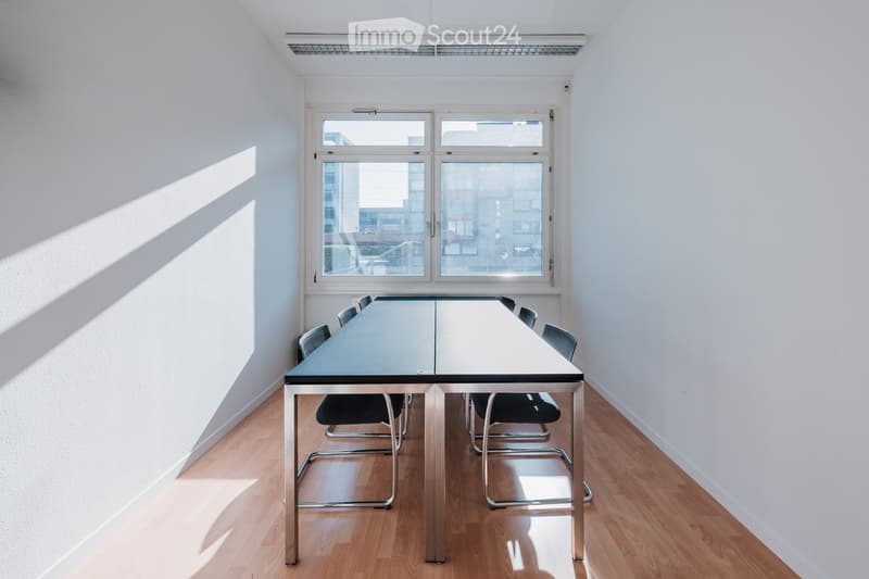 Bright, minimalist room with a long conference table and chairs, large windows, and wooden flooring.