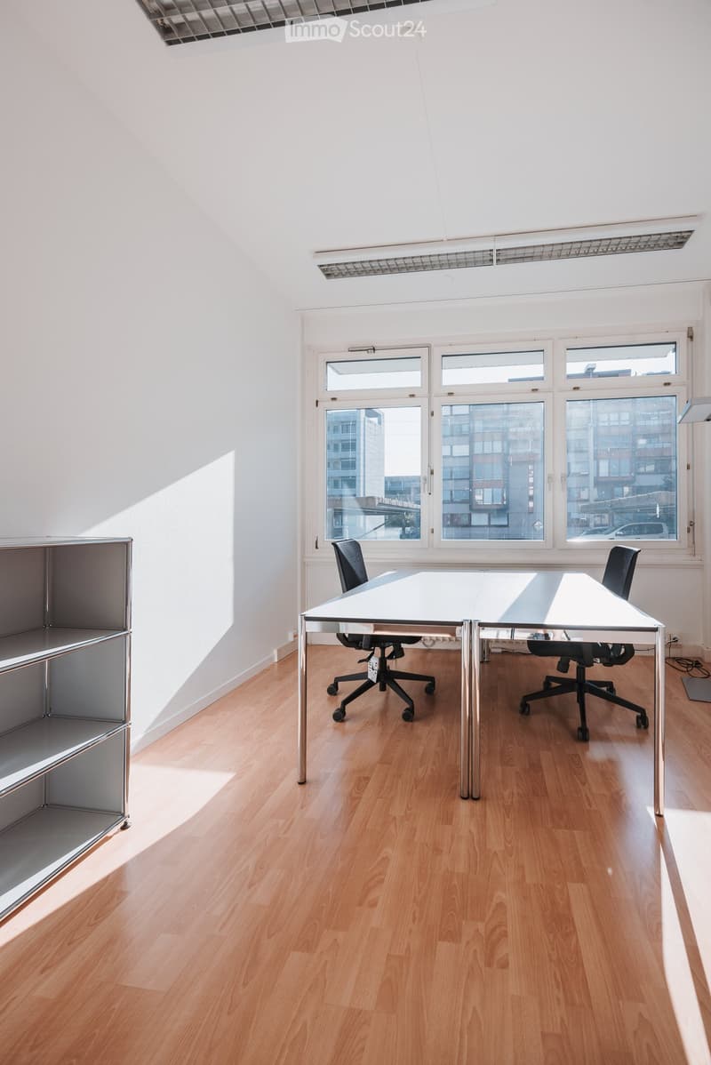 Bright, minimalist room with wooden flooring, large windows, a white table, two black office chairs, and a metal shelf unit.