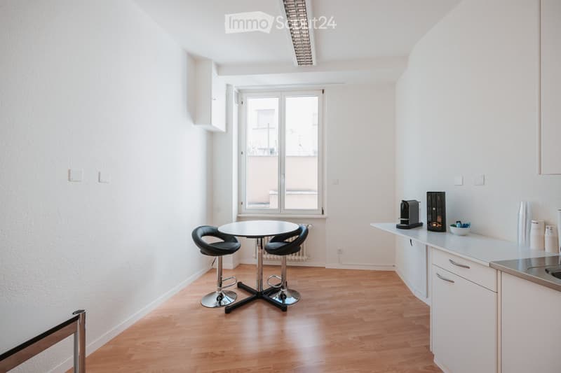 A modern, open-plan living and dining area with a round table and two stools, a kitchen counter with a coffee maker, and white cabinets.