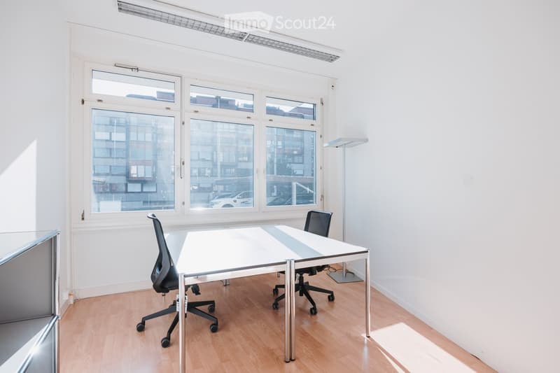 Bright, minimalist office space with large windows, a white table, and two black office chairs.
