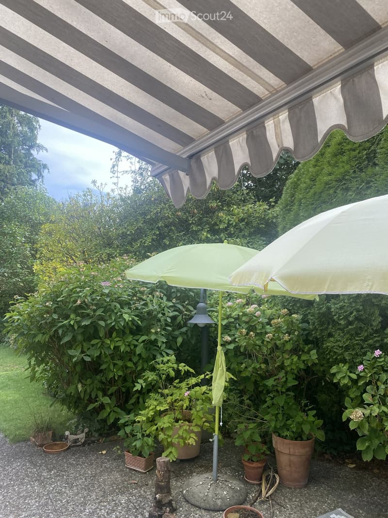 A shaded outdoor area with a striped awning, potted plants, and two large umbrellas, surrounded by lush greenery and a gravel ground.
