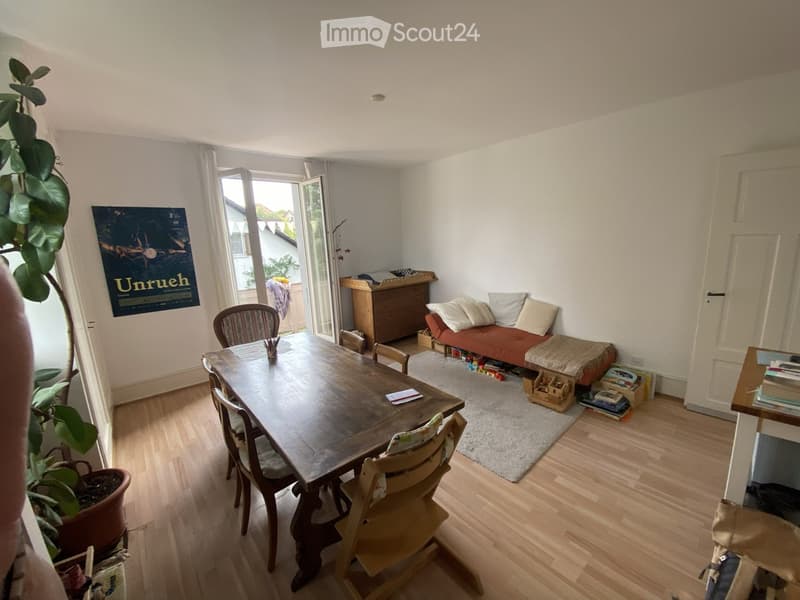 A well-lit living and dining area with a wooden dining table and chairs, a small sofa, a potted plant, and a poster on the wall. The space features light wooden flooring and large windows with white curtains.