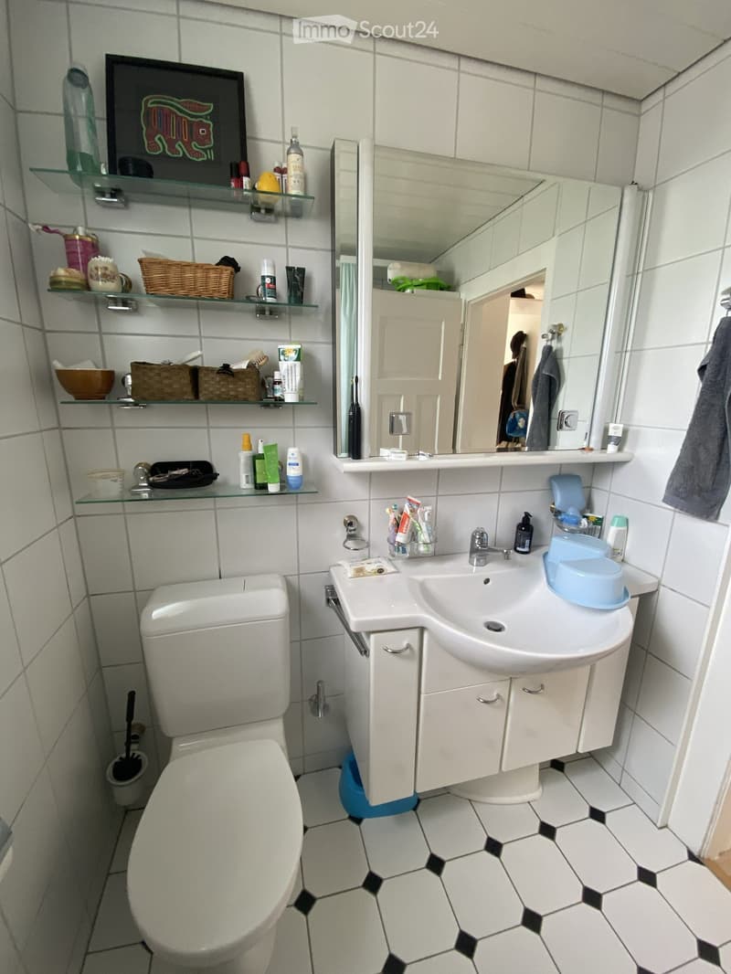 A small bathroom with white tiles, a toilet, a sink with a mirror, glass shelves holding various toiletries, and a wicker basket on the shelves.