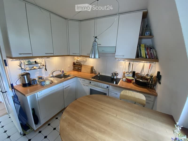 Modern kitchen with white cabinets, wooden countertops, and various appliances including a sink, oven, dishwasher, and coffee maker. A small dining area with a wooden table and chair is visible.