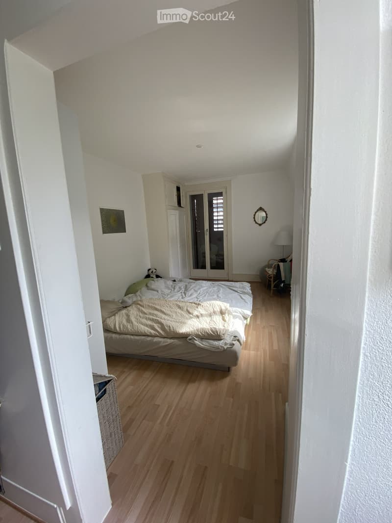 Bedroom with wooden flooring, a bed with beige bedding, a wardrobe, a mirror, a lamp, and a small chair.