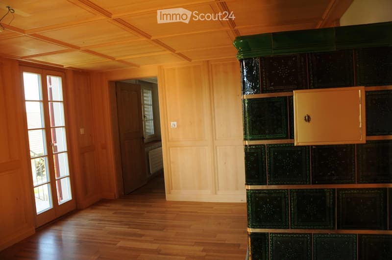 Bright, wooden-floored room with a large, decorative green and black fireplace, large windows, and a door leading to another room.