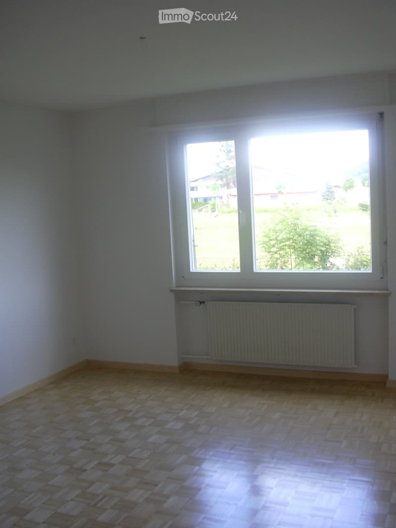 Empty room with wooden flooring, white walls, and large windows with a radiator below them.