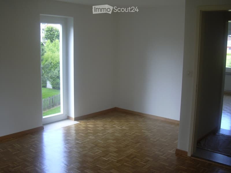 Empty room with wooden parquet flooring and white walls, featuring a large window with a view of a green garden.