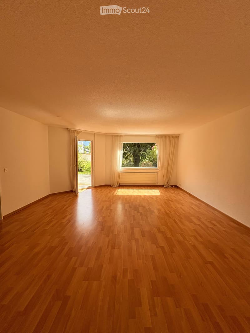Empty room with wooden flooring, large windows with curtains, and a sliding door leading to an outdoor area.