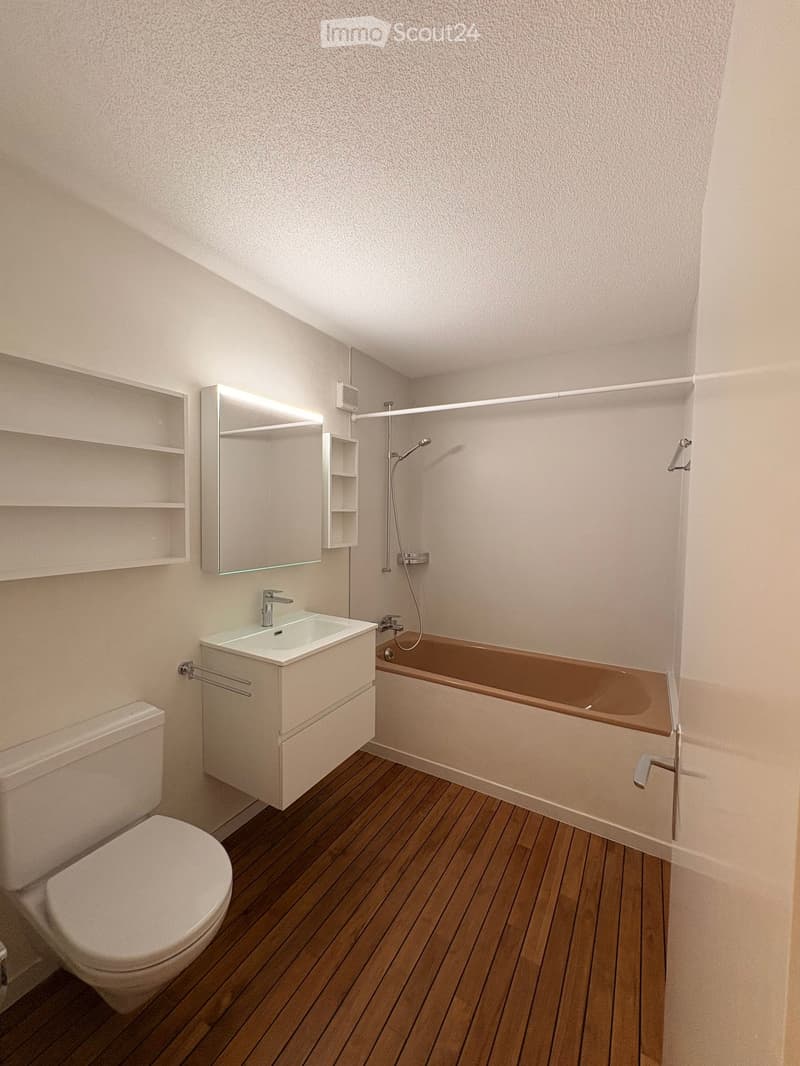 Modern bathroom with a wooden floor, white walls, a bathtub, a sink, a toilet, a mirror, and a shower. The sink has a modern faucet, and the mirror is illuminated.