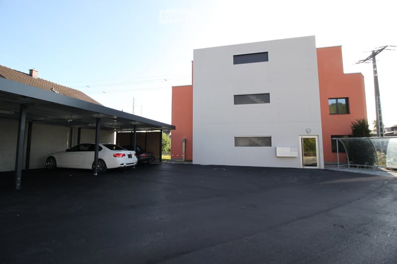 Modern two-story building with a covered parking area. The building has a white facade with orange accents and several windows. Two cars are parked under the covered area.