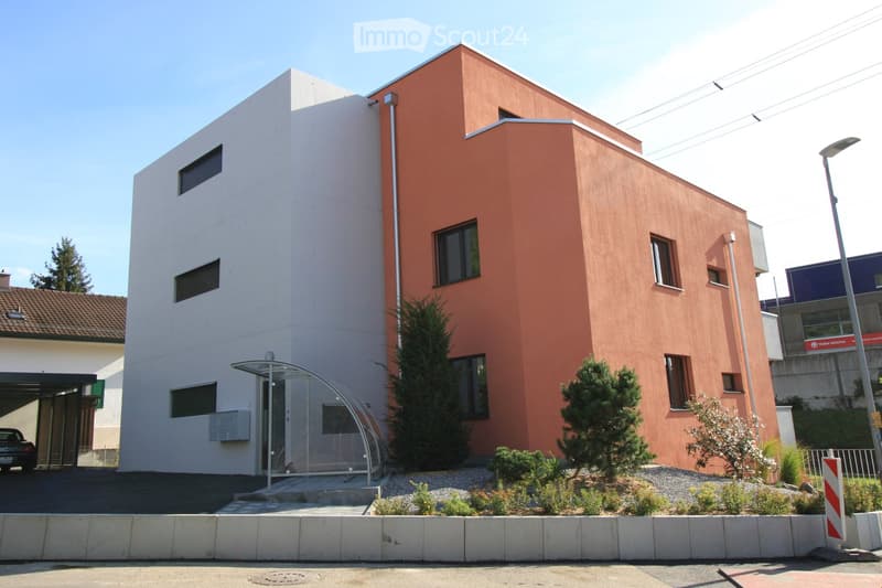 Modern two-story building with a combination of white and orange facades, featuring multiple windows and a small entrance canopy. There is a small garden area with shrubs and trees in front, and a car is parked in a nearby garage.