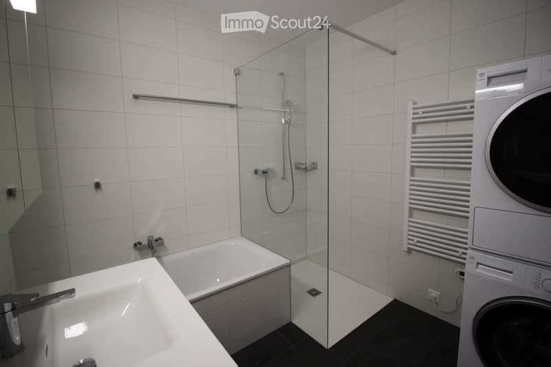Modern bathroom with white tiles, a bathtub, a glass-enclosed shower, a sink, a towel rack, and a washing machine.
