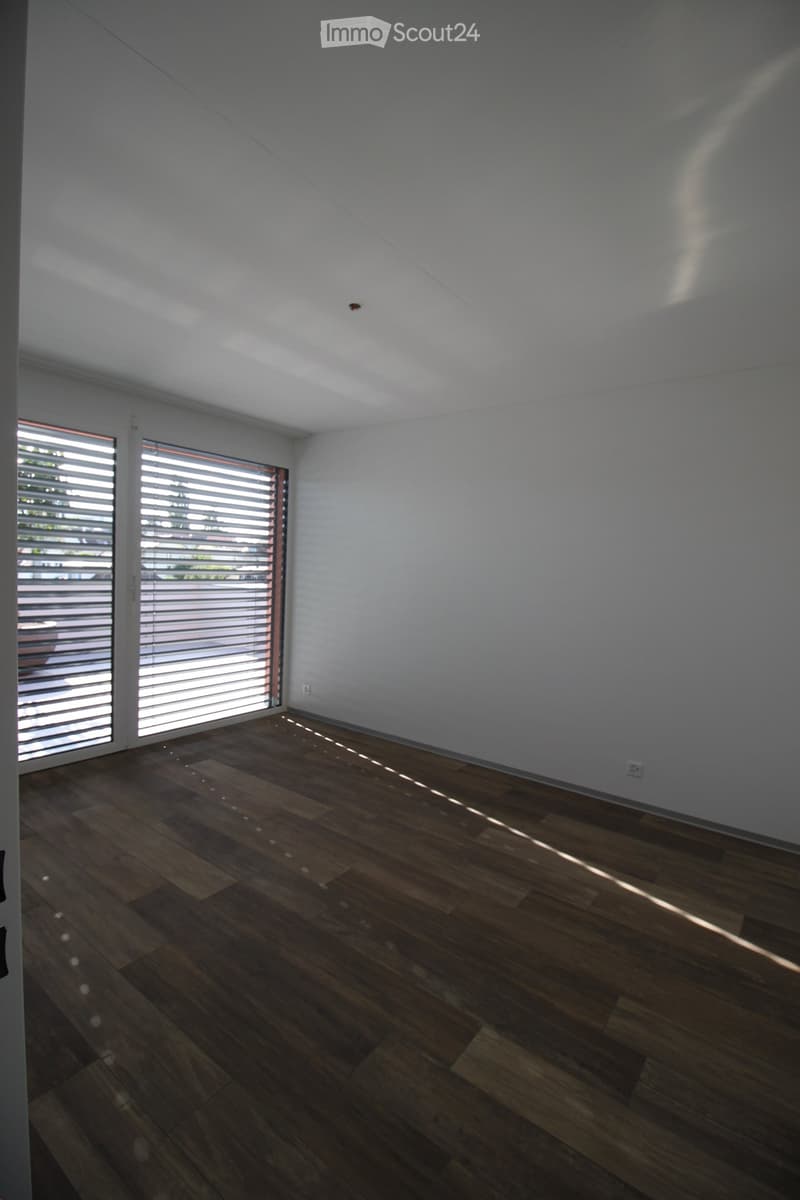 Empty room with wooden flooring and large windows with blinds, allowing natural light to enter.