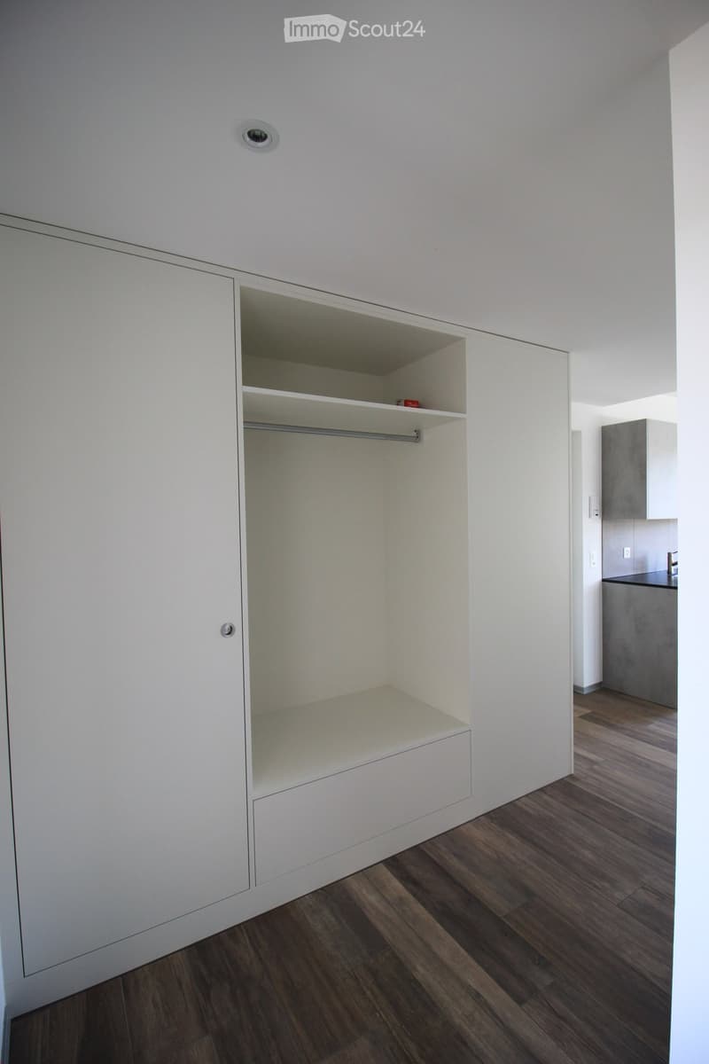 Empty room with white built-in closet and wooden flooring, adjacent to a small kitchen area.