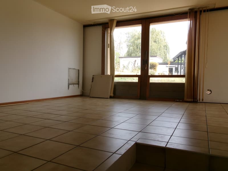 Empty room with tiled floor, large sliding glass door leading to outdoor area, and a small piece of furniture leaning against the wall.