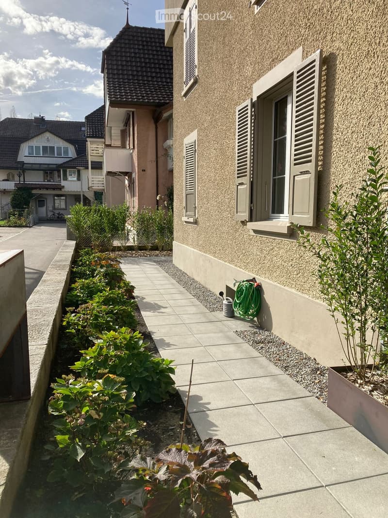A narrow garden area with a paved pathway, green plants, and a hose coiled against the building wall.