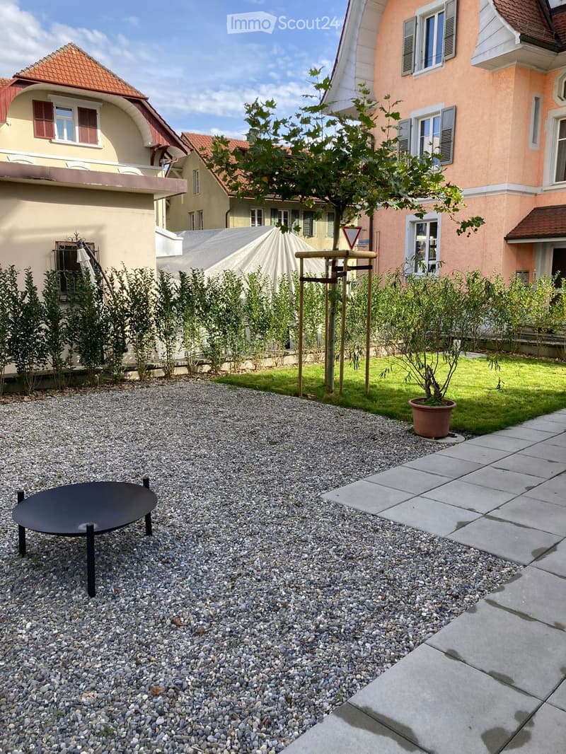 Garden area with gravel, a round black table, a potted plant, and a small wooden structure, surrounded by houses.
