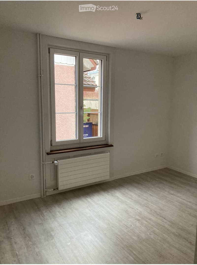 Empty room with light wooden flooring, white walls, and a window with a radiator below it.