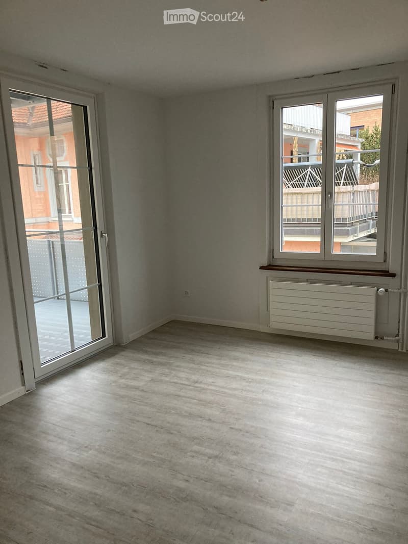 Empty room with light grey walls, concrete floor, large window, and sliding glass door leading to a balcony.