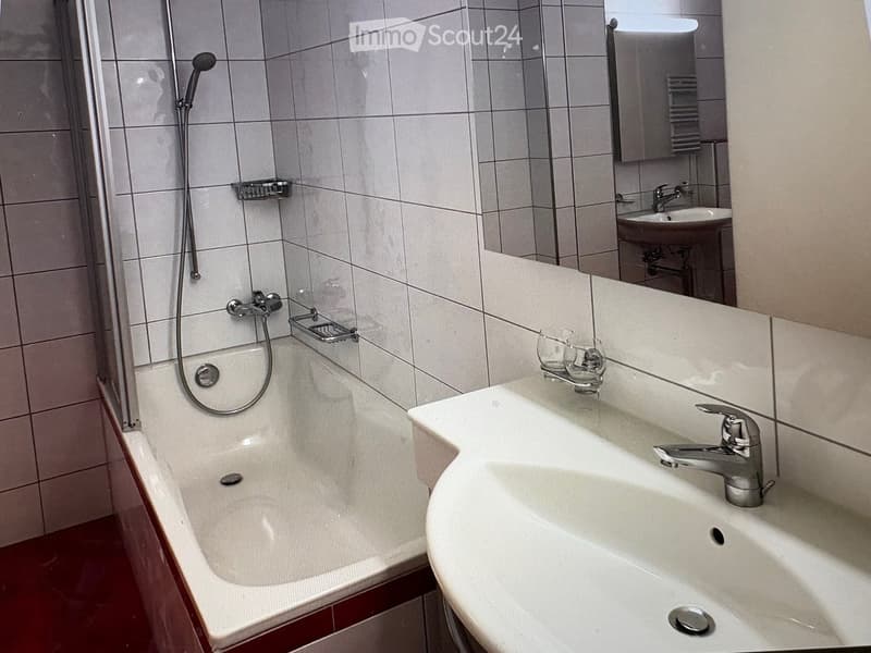 Modern bathroom with a bathtub, sink, mirror, and showerhead. The walls are tiled, and there are two glasses on the sink counter.