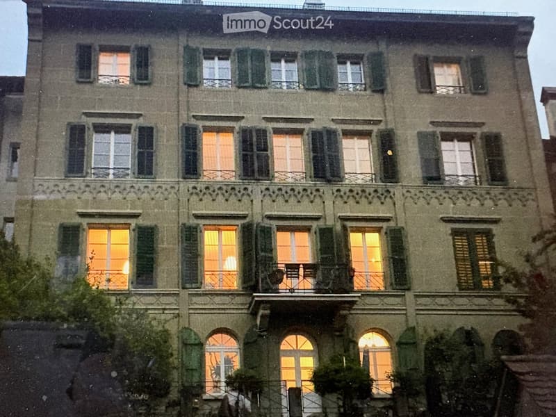 A multi-story residential building with multiple windows, some illuminated from within, and decorative balconies. The facade is made of stone with green shutters on the windows.