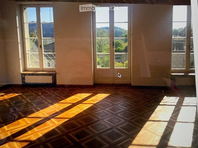 Empty room with wooden floor and large windows, allowing natural light to fill the space. The room has a simple layout with no furniture or decorations.