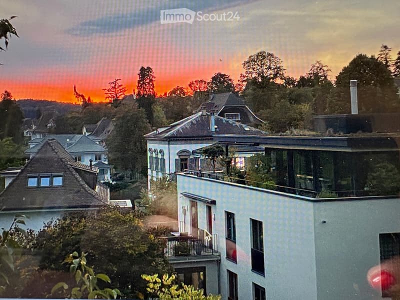 A multi-story house with a modern design, featuring large windows, a balcony, and a flat roof, set against a scenic hillside backdrop with trees and a dramatic sunset sky.