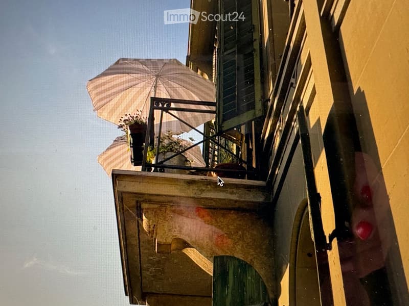 Balcony with wooden railing, green shutters, and a large umbrella providing shade. Potted plants are visible on the balcony.