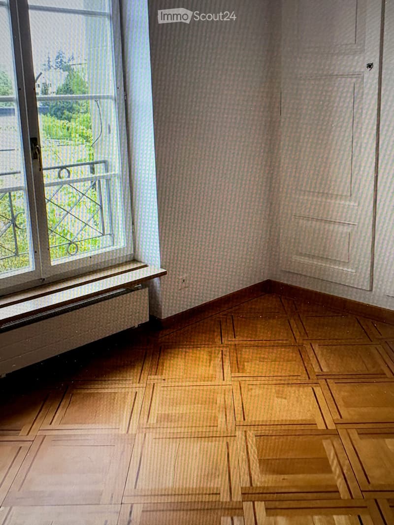 Empty room with wooden flooring and patterned wallpaper, featuring a window with a radiator below and a metal security grate.