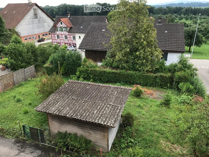 Aerial view of a rural residential area with multiple houses, surrounded by greenery and trees. Houses include a traditional half-timbered house, a modern white house, and a wooden shed. The area is lush with vegetation and includes a wooden fence and a s