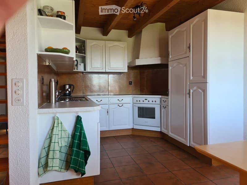 A cozy kitchen with white cabinets, a stainless steel sink, a white oven, and a refrigerator. Green checkered towels hang on the counter, and a small table is visible in the foreground.