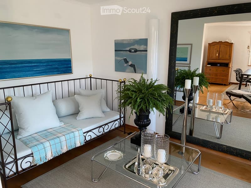 Elegant living room with a white metal-framed sofa, plaid throw, glass coffee table, decorative candle holders, potted plant, and a large mirror reflecting a dining area with a wooden cabinet and chairs.