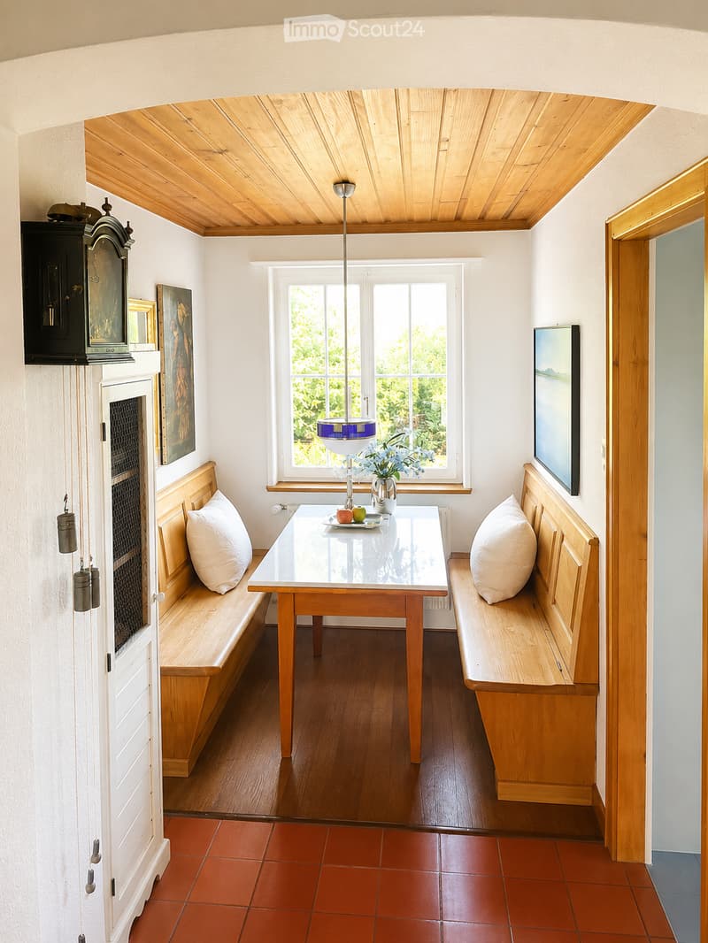 Bright kitchen with wooden benches, a glass table, pendant light, and a vase with flowers on the table. The room features a slanted wooden ceiling and large windows with a view of greenery.