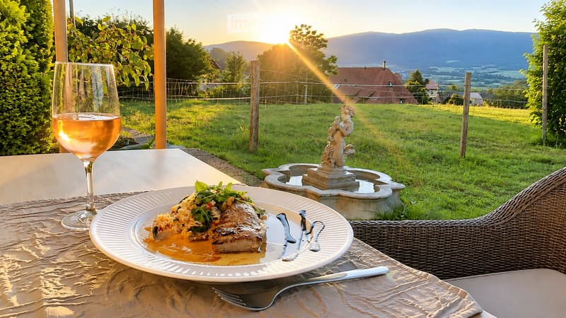 Outdoor dining setup with a plate of food, wine glass, and a scenic view of a valley and mountains at sunset.