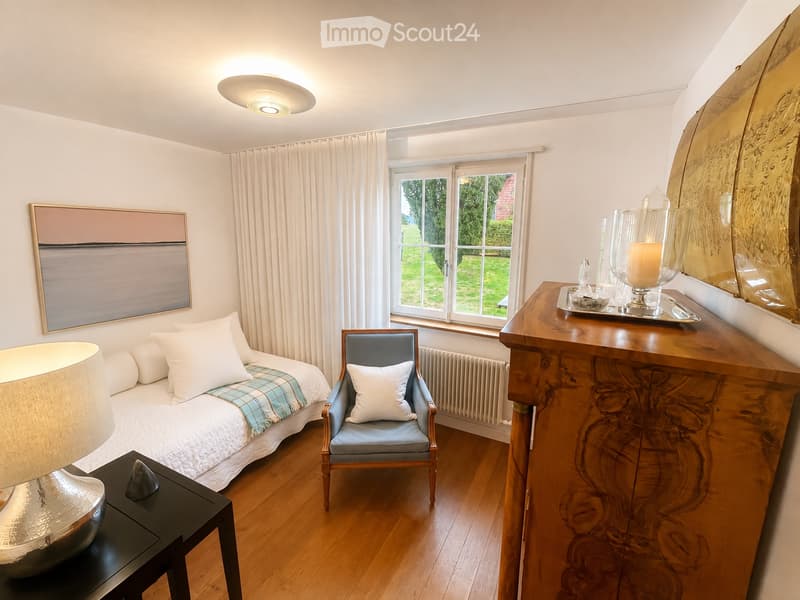 Cozy bedroom with a single bed, a wooden chair, a nightstand with a lamp, a large mirror, and a decorative tray with candles. The room features wooden flooring, white walls, and large windows with white curtains.