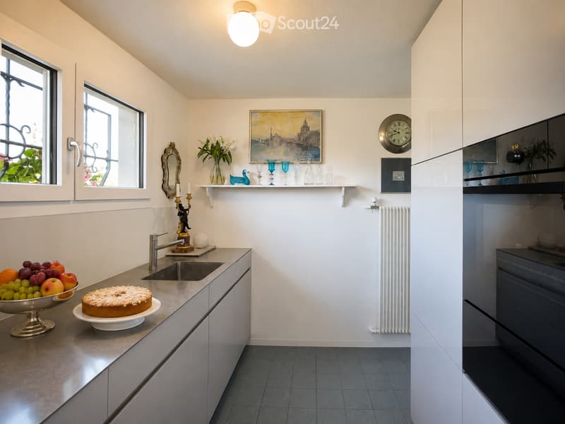 Modern kitchen with white walls, grey cabinets, and a marble countertop. Features a sink, fruit bowl, cake, mirror, clock, radiator, and a refrigerator.