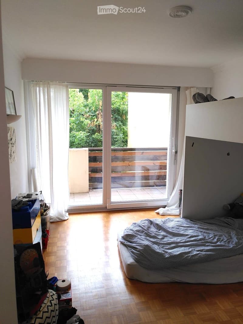 Bright bedroom with wooden flooring, large sliding glass doors leading to a balcony, a bed with striped bedding, a nightstand, and a mirror on the wall.