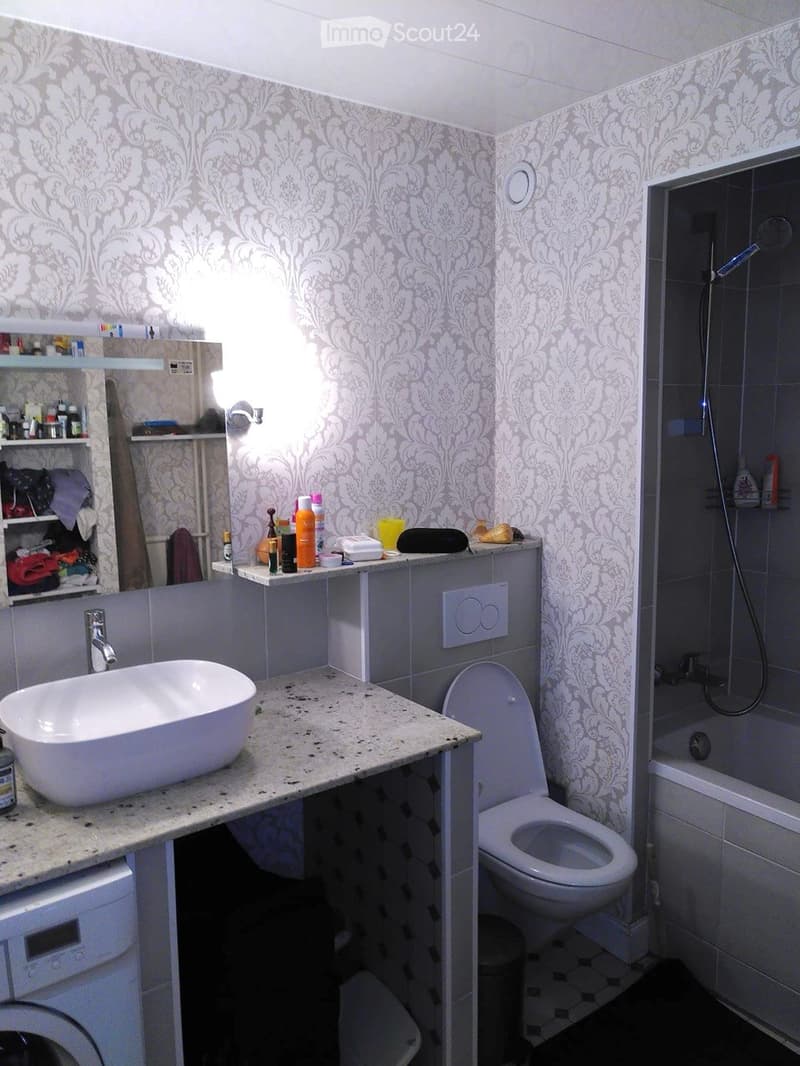 Bathroom with a sink, toilet, bathtub, and shower. The walls are covered with patterned wallpaper, and there is a shelf with various toiletries and a washing machine visible.