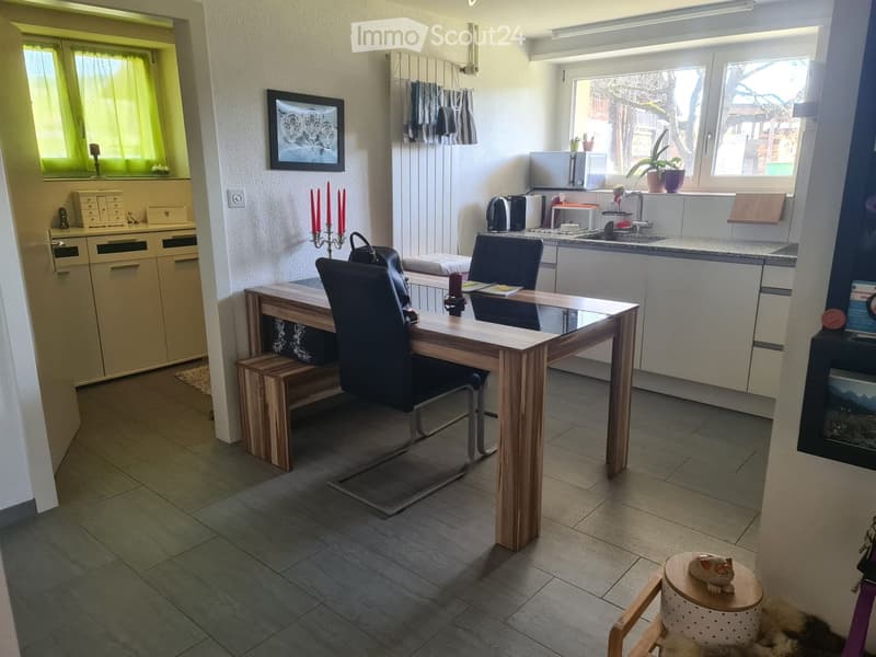 A modern, well-lit room with a kitchen area and a dining area. The kitchen features white cabinets, a sink, a microwave, and a faucet. The dining area includes a wooden table with two black chairs and a small bench. A small stool with a decorative item si