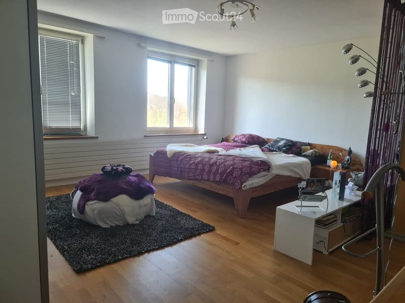 Bright bedroom with wooden flooring, large windows, a double bed with purple bedding, a nightstand, a small table with a lamp, and a decorative rug.