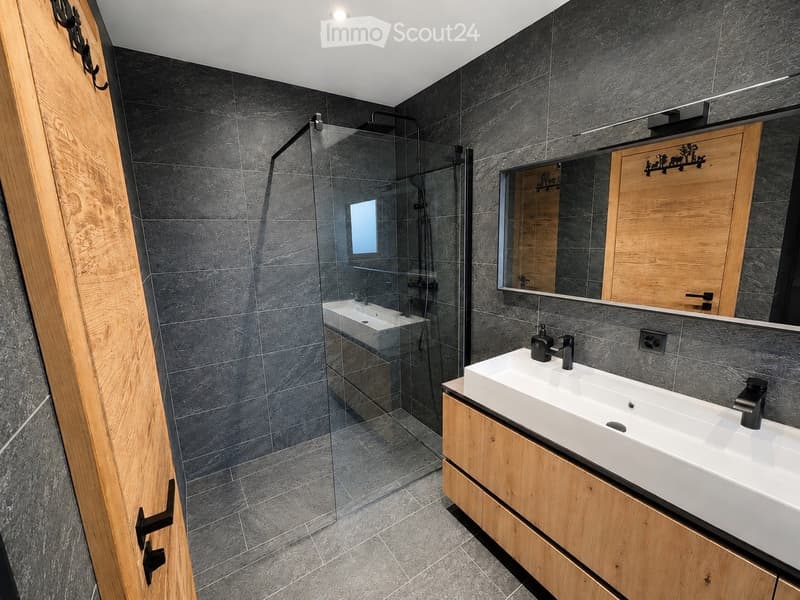 Modern bathroom with dark grey tiles, a glass-enclosed shower, double sink vanity with wooden cabinets, and a large mirror.